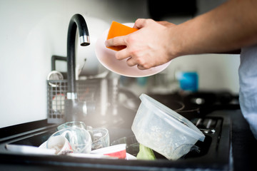 The man washes the dirty dishes in the kitchen by hand. The man holds a dirty plate and a washing sponge in his hand. The concept of washing dirty dishes by hand.