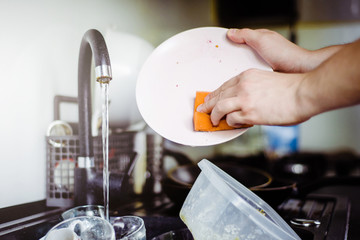 The man washes the dirty dishes in the kitchen by hand. The man holds a dirty plate and a washing sponge in his hand. The concept of washing dirty dishes by hand.