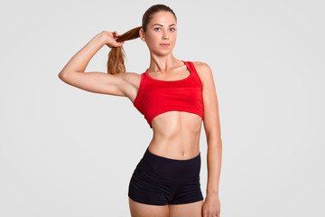 Fototapeta na wymiar Horizontal shot of slim sporty woman has nice healthy muscular body, dressed in casual top and shorts, touches pony tail, poses against white background. Athletic girl takes break after workout