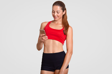 Fototapeta na wymiar Studio shot of pleased fit female takes break after cardio training, holds up to date cell phone, surfes social networks, reads news about sport in internet, dressed in casual top and shorts