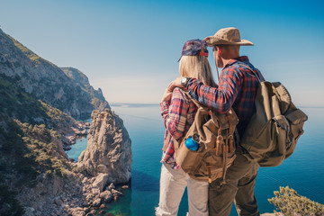 Man and woman backpackers standing and hugging on the mountain. Couple hikers with backpacks relaxing on top of a hill and enjoying sea view and cliffs. - obrazy, fototapety, plakaty
