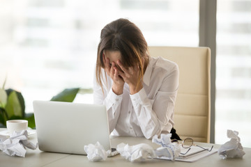 Stressed tired businesswoman feels exhausted sitting at office desk with laptop and crumpled paper,...