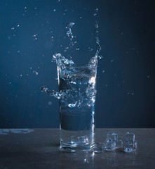 Ice splashes into glass of water on blue background
