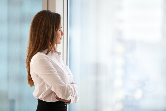 Successful thoughtful woman business leader looking out of big window enjoying city view and wellbeing feeling motivated for new goals, rich millennial businesswoman dreaming of future, copy space