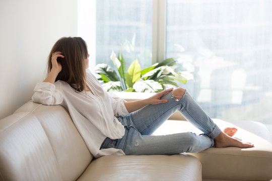 Relaxed thoughtful woman looking out of big window enjoying view and wellbeing dreaming at luxury living room, rich millennial lady taking pleasure of stress free weekend resting on comfortable sofa