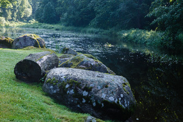a green, natural landscape with a slowly flowing river with many water plants in it, with a riverbank with large stones and a wooden block in the foreground; tree and grass reflection in the water