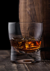 Glass of whiskey with ice cubes next to wooden barrel. Cognac brandy drink