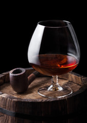 Glass of cognac brandy drink and vintage smoking pipe on top of wooden barrel.