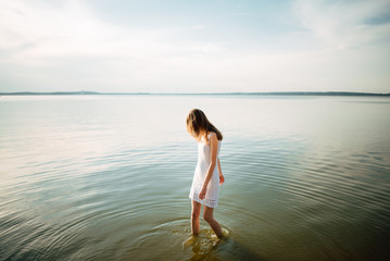 Woman in summer dress standing on seashore and looking at horizon. Young beautiful girl standing in water