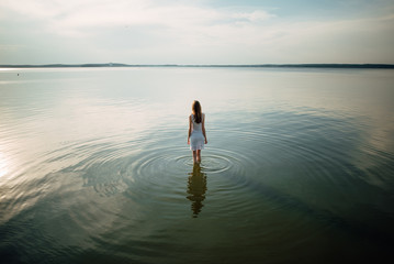 Woman in summer dress standing on seashore and looking at horizon. Young beautiful girl standing in water - Powered by Adobe