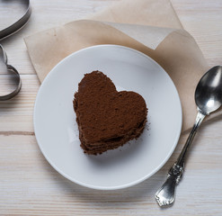 top view of a chocolate heart-shaped dessert lying on a saucer next to a silver spoon and iron molds on a white wooden background