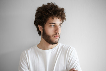 Close up shot of attractive stylish young unshaven man with fuzzy beard and voluminous hair having serious look, posing at white blank studio wall. Human facial expressions, emotions and feelings