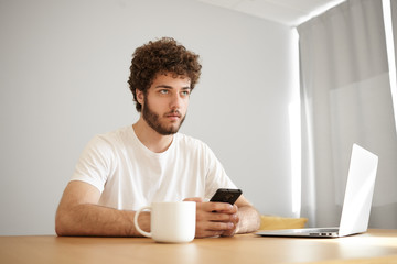 Fashionable young unshaven guy with wavy hair having pensive look typing sms using wifi on smart phone, browsing internet on portable computer and drinking coffee. People, lifestyle and technology