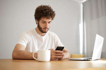 Candid shot of focused young bearded male freelancer in white t-shirt typing text message on mobile phone while working distantly using laptop and drinking coffee, sitting at his workplace