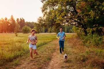Fototapeta premium Young couple walking pug dog in autumn forest. Happy puppy running along and having fun playing with master.