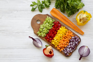 Chopped fresh vegetables (carrot, celery, onion, colored peppers) arranged on cutting board on white wooden table, view from above. Overhead, top view.