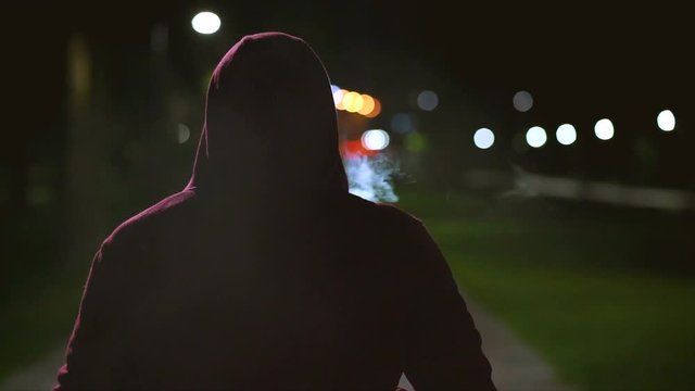The man in a hood walking and smoking on the night street. slow motion