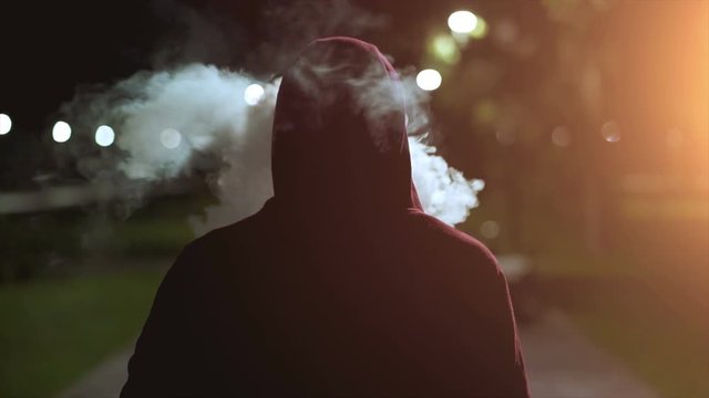 The fellow in a hood walking and smoking on the night street. slow motion