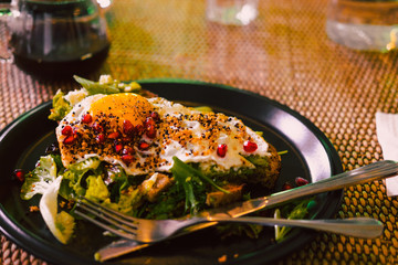 Avocado toast with egg and pomegrate seeds on black plate at restaurant table. Dark food photography concept. Copy space