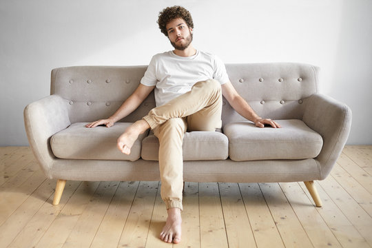 Casually dressed young European guy with bare feet and fuzzy beard relaxing in living room, sitting on sofa, keeping legs crossed. People, lifestyle, leisure, style, interior and fashion concept