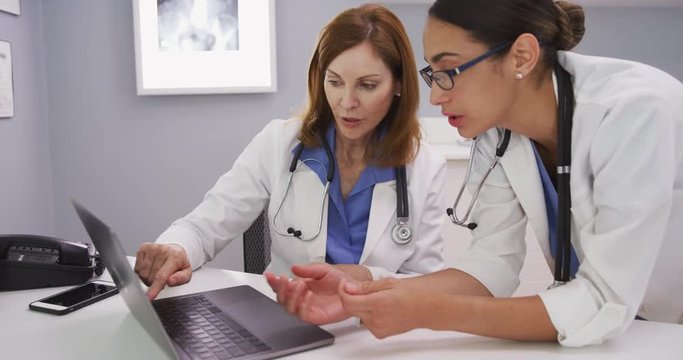 Young latina doctor looking at laptop computer and talking with elder physician