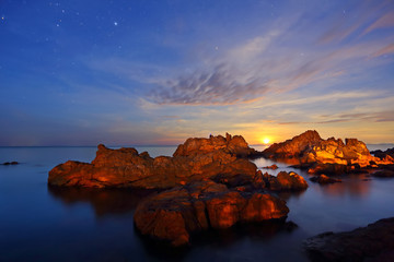 Long Exposure of stars, the moon is falling with rocks and sea in the dawn