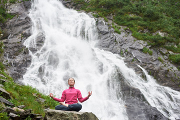 Naklejka na ściany i meble Young female practising yoga, meditating, relaxing alone against waterfall Balea in Fagarash mountains. Rocky waterfall in the mountains.