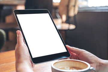 Mockup image of hands holding black tablet pc with blank white screen with coffee cup on wooden...
