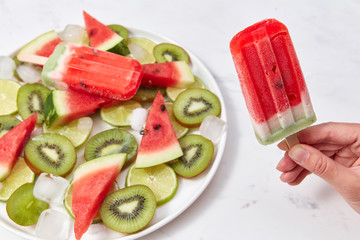 A woman's hand holds an ice cream on a stick in the form of a piece of watermelon on a background of a plate with pieces of different fruits around a white background with space for text.