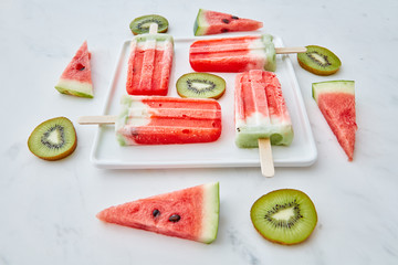 Slices of watermelon and kiwi in the form of a circle and a triangle around a plate with a healthy ice cream lolly in the shape of a square on a gray marble background . Food pattern.