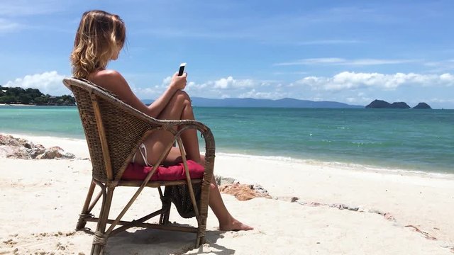 The young beautiful woman sits on a chair by the sea with a phone in hands.