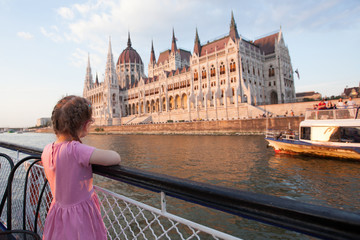 A little girl in red glasses sails on a ship on the Danube and looks at the parliament building in...