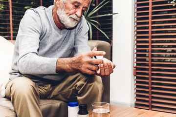 Senior man holding bottle with pills and taking medicine with glass of water sitting on sofa in living room at home. Healthcare senior people concept