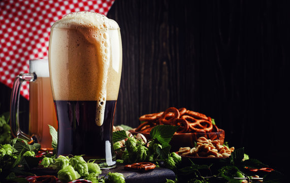 Dark German beer is poured into a glass, fresh green hops and bowls with salty snacks and nuts, autumn beer festival concept, dark background, selective focus