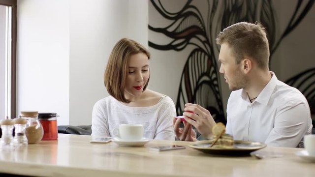 Two young people is sitting in a cafe. Man and woman are smiling and sitting in the cafe at the table during break. Couple is talking and drinking coffee. He proposes marriage