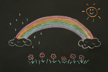 Actual drawing on black chalk board. Colorful rainbow on clouds with sun shining it's light,  some...