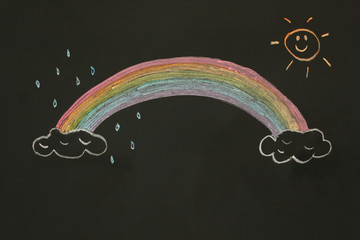 Actual drawing on black chalk board. Colorful rainbow on clouds with cute sun shines it's light and...