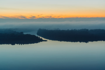 Fototapeta na wymiar Broad river flows along shore with forest under thick fog. Early blue sky reflected in water. Yellow glow in picturesque predawn sky. Colorful morning mystical atmospheric landscape of majestic nature