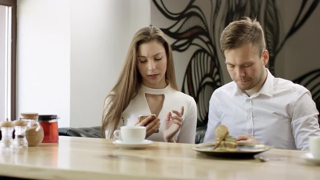 Two young people is sitting in a cafe. Man and woman are smiling and sitting in the cafe at the table during break. Couple is talking and drinking coffee. They aretyping mesagies on their moile phones