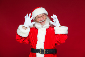 Fototapeta na wymiar Christmas. Good Santa Claus in white gloves shows faces, grimaces, shows his tongue. Not standard behavior. Isolated on red background.