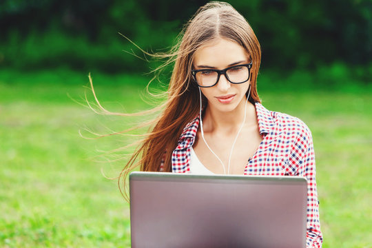 Portrait of young cute woman in plaid shirt wearing glasses using laptop in the park sitting on the green grass. Leisure time activity concept