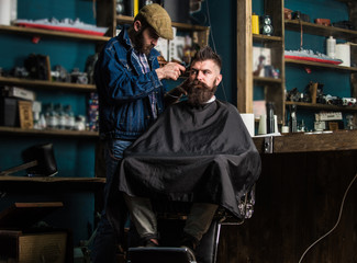 Barber with clipper trimming hair on temple of bearded client. Hipster client getting haircut. Barber with hair clipper work on hairstyle for hipster, barbershop background. Hipster lifestyle concept