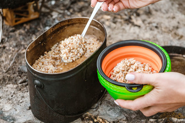 A woman camper puts food from a cauldron in a plate, cooking during a hike