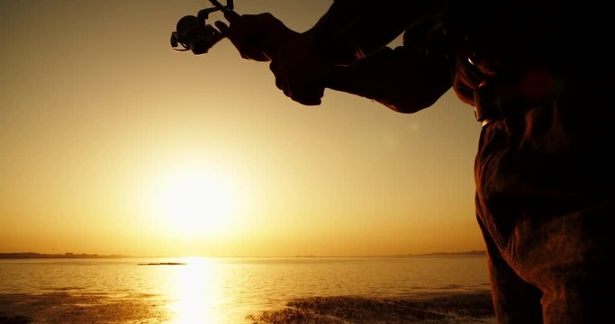 An experienced Fishman catching fish with professional spinning rod from a lake shore. lake in orange sunset 4k