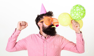 Celebration concept. Guy in party hat with air balloons celebrates. Hipster in giant sunglasses celebrating birthday. Man with beard and mustache on busy face blows into party horn, white background