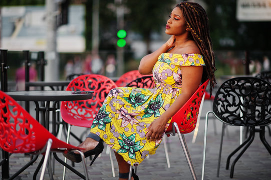 Cute small height african american girl with dreadlocks, wear at coloured yellow dress, sitting at outdoor cafe on red chair.