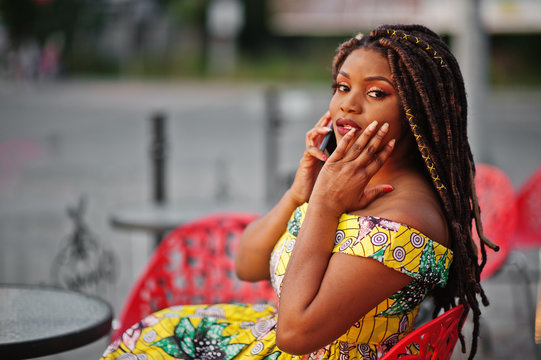Cute small height african american girl with dreadlocks, wear at coloured yellow dress, sitting at outdoor cafe on red chair and speaking on phone.