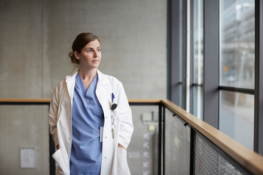 Thoughtful female doctor standing with hands in pockets while looking through window at hospital corridor
