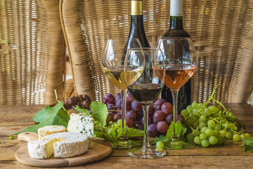 French food and drink/Two bottle of wine and three glasses of white, pink and red wines, grapes, various of french cheeses on vintage wooden table with wicker chairs