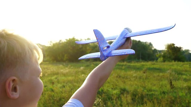 Closeup back view of cute white kid holding blue toy plane in hand outside in sunny sunset green meadow. Boy imagines toy to be flying. Travel concept. Real time 4 k video footage.